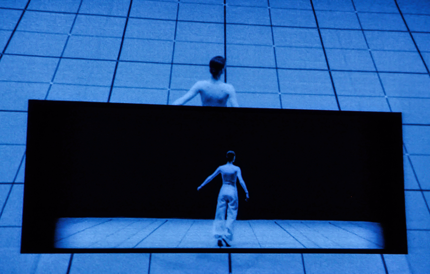 A film still of one dancer with their back to the audience against a grid of black lines. Below them is a dancer in a black square, also with their back to the camera.